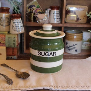May include: A cylindrical ceramic sugar canister featuring alternating horizontal olive green and cream white stripes with the word SUGAR printed in black lettering on the front, accompanied by a matching lid with a small knob handle.