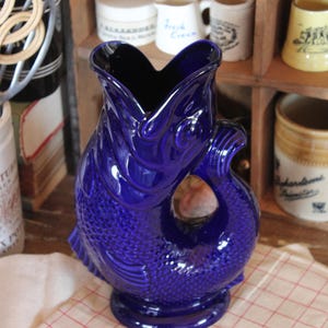 May include: A cobalt blue ceramic pitcher with a fish design. The pitcher has a wide, open mouth and a handle that forms a loop. The fish scales are textured. The pitcher is on a red and white checkered tablecloth.