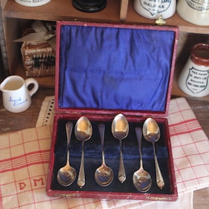 Può includere: Un set di sei cucchiai d'argento esposti in una scatola rossa foderata in velluto blu. I cucchiai hanno un design classico. La scatola è aperta, rivelando i cucchiai contro il tessuto blu. Altri oggetti vintage sullo sfondo.