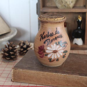 May include: A tan ceramic jar with a cork stopper, featuring hand-painted floral designs in white, green, and dark red. The text "Herbes de Provence" is written in black script on the front of the jar, which has a rounded body and a flared rim.