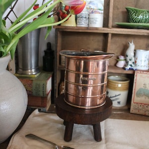 May include: A copper creamer with the word "CREAMER" stamped on the side, sitting on a small wooden stool. A silver spoon rests on a white cloth. Pink and white tulips are in a vase.