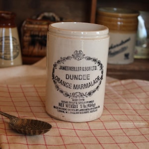 May include: A vintage white ceramic jar of Dundee Orange Marmalade with a black floral label and text. The jar is 4.5 inches tall and sits on a red and white checkered cloth. A decorative spoon rests nearby.