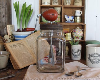 Könnte beinhalten: Vintage Glas-Butterfass mit einer roten und silbernen Handkurbel. Das Fass steht auf einem Tisch mit einem Buch und anderen Vintage-Gegenständen.