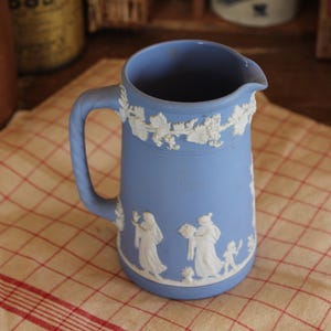 May include: A light blue ceramic pitcher with a handle and spout. The pitcher features white relief designs of figures and floral patterns around the rim. It sits on a red and white checkered cloth.