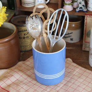 May include: A light blue ceramic utensil holder with two white stripes, holding a silver fork, a whisk, and a woven utensil. The holder sits on a red and white checkered cloth, with other vintage kitchen items in the background.