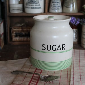 May include: Cream-colored ceramic sugar jar with a lid, green stripes, and the word "SUGAR" in black. A silver spoon rests on a red and white checkered cloth. Other vintage jars and containers are in the background.