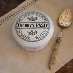 May include: A white ceramic lidded pot with black text reading ANCHOVY PASTE, AN EXCELLENT RELISH FOR BREAKFAST LUNCHEON & C, featuring small thistle illustrations. Beside it lies a metal spoon with an ornate bowl and a ribbed handle.
