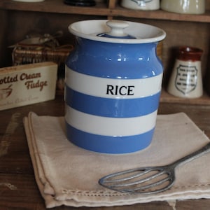 May include: A blue and white striped ceramic rice jar with a lid, the word "RICE" is printed on the front. It sits on a beige linen napkin with a metal whisk. The jar is approximately 15 cm tall.