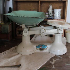 May include: A vintage white kitchen scale with a mint green bowl and a round platform. The scale has a classic design with a logo on the base. A spoon and a decorative cloth are in the foreground.