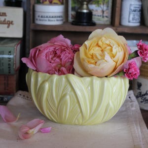Pode incluir: Uma tigela de cerâmica amarelo pálido com um desenho de folhas, cheia de rosas rosa e amarelas e pequenas flores rosa. A tigela está sobre um pano branco, com pétalas caídas por perto. O fundo inclui itens vintage.