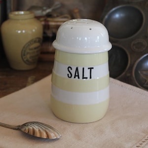 May include: A vintage-style salt shaker with a white lid and a cylindrical body. The body is pale yellow with horizontal white stripes and the word "SALT" in black. A decorative spoon rests on a linen napkin.