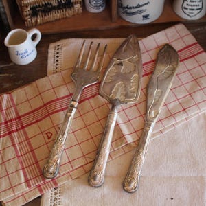 Pode incluir: Três utensílios de servir de prata antigos: um garfo, uma espátula para bolo e uma faca, dispostos sobre um guardanapo de linho xadrez vermelho e branco. Os utensílios têm cabos ornamentados. Uma pequena cremeira branca com "Fresh Cream" é visível.