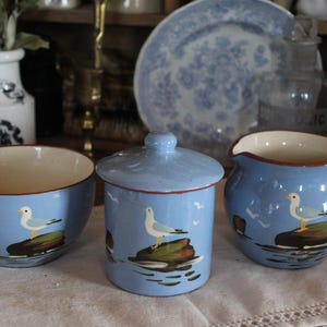 Könnte beinhalten: Ein Set aus drei blauen und weißen Keramik-Küchenutensilien mit einem Möwen-Design. Das Set beinhaltet eine Zuckerdose mit Deckel, eine Milchkännchen und eine kleine Schüssel. Jedes Stück zeigt eine Möwe, die auf einem Felsen sitzt, mit einem blauen und weißen Meereshintergrund.