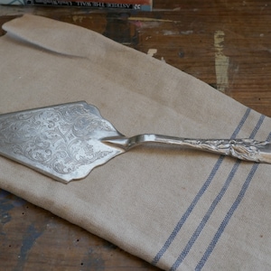 May include: Silver cake server with ornate floral engraving on the blade and handle. The server rests on a beige linen cloth with blue stripes. A vintage kitchen utensil for serving cakes and pies.