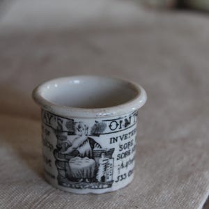 May include: Small, white ceramic ointment pot with black printed text and illustrations. The pot has a rounded rim and a cylindrical body. The text includes the word "OINT" and other illegible text. The pot is sitting on a light-colored surface.