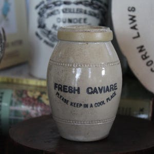 May include: A white ceramic jar with a black label that reads "FRESH CAVIARE PLEASE KEEP IN A COOL PLACE".