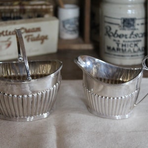 Może przedstawiać: Srebrny zestaw do śmietanki i cukru. Cukiernica ma uchwyt i uchwyt na zawiasach. Śmietanka ma zakrzywioną wylewkę i ozdobny uchwyt. Oba elementy mają żebrowany wzór i są umieszczone na jasnej powierzchni.