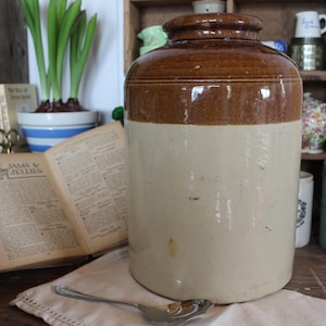 Può includere: Un grande vaso di ceramica marrone e bianco con un'ampia apertura. Il vaso è appoggiato su un tavolo di legno con un libro e un cucchiaio.