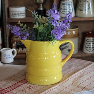 Może przedstawiać: Jasnożółty ceramiczny dzbanek z uchwytem, wypełniony fioletowymi kwiatami i zielenią. Dzbanek ma poziome żłobienia i stoi na czerwono-białej kraciastej szmatce. Zabytkowe słoiki i pojemniki w tle.