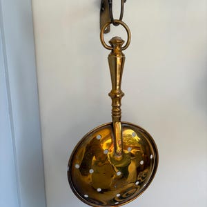 May include: A polished brass skimmer with a long, tapered handle and a circular bowl featuring a series of evenly spaced drainage holes. The utensil is suspended from a curved brass hook attached to a wall-mounted bracket, showcasing a reflective metallic finish.
