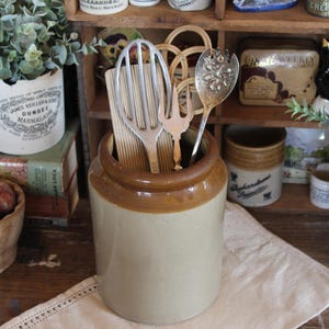 May include: A stoneware kitchen crock with a brown rim holds various vintage utensils, including a whisk, forks, and a spoon. The crock sits on a cream-colored linen cloth, with other antique jars and a wooden shelf in the background.