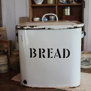 Peut inclure: Boîte à pain rectangulaire blanche avec couvercle et poignée. Le mot "BREAD" est inscrit en noir sur le devant. La boîte a un aspect vintage et vieilli, avec des signes d'usure visibles.