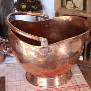 Puede incluir: Un cubo para carbón grande y redondo de cobre martillado con una parte superior ancha y abierta y un asa de metal arqueada y resistente unida a los lados. El recipiente presenta una superficie metálica pulida y reflectante.