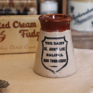 Puede incluir: Una pequeña jarra de crema antigua con borde marrón y cuerpo beige. La jarra tiene una etiqueta negra con el texto "THE DAIRY ST. JOHN'S LANE HALIFAX RICH THICK CREAM". Una caja de fudge de crema cuajada está en el fondo.