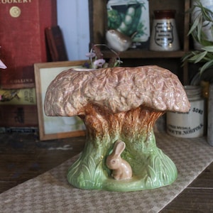 Peut inclure: Un pot en céramique en forme d'arbre avec une petite figurine de lapin assise dans la base. Le pot est peint dans des tons de brun, vert et rose.
