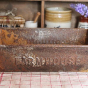 Puede incluir: Una bandeja de pan rectangular de metal marrón oxidado con la palabra "FARMHOUSE" en relieve en la parte delantera. La bandeja está sobre un paño a cuadros rojos y blancos. Otros artículos de cocina vintage están en el fondo.