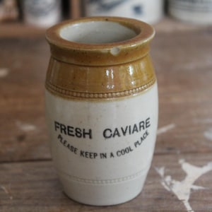 Op de afbeelding: Een vintage keramische kaviaarpot met een bruine rand en accenten, en een witte body. De pot heeft de woorden "FRESH CAVIARE" en "PLEASE KEEP IN A COOL PLACE" in zwarte letters. De pot is cilindrisch met een licht afgeronde basis.