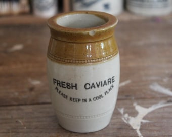 May include: A vintage ceramic caviar jar with a tan rim and accents, and a white body. The jar has the words "FRESH CAVIARE" and "PLEASE KEEP IN A COOL PLACE" printed in black. The jar is cylindrical with a slightly rounded base.
