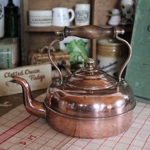 Puede incluir: Una tetera de cobre con asa y boquilla de madera. La tetera está sobre un paño a cuadros rojos y blancos. La imagen también incluye un libro de cocina y otros artículos de cocina vintage.