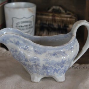 May include: A ceramic gravy boat featuring a white base with a light blue floral pattern. The vessel has a scalloped rim, a curved handle, and stands on three small feet. The design includes intricate blue flowers and foliage across the exterior.