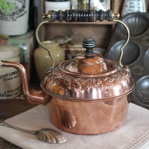May include: A copper tea kettle with a decorative floral pattern around the top and a black and wood handle. The spout is curved and the kettle sits on a white cloth. A vintage kitchenware item.