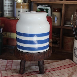 Peut inclure: Un pot en céramique blanche avec des rayures bleues posé sur un support en bois à trois pieds. Le pot a un couvercle et est décoré de rayures bleues et blanches. Le pot est posé sur une table avec une nappe à carreaux rouge et blanc.