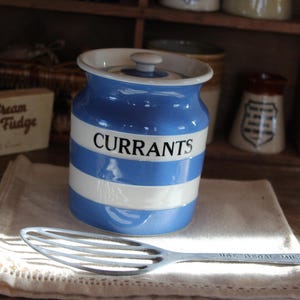 May include: A blue and white striped ceramic jar labeled "CURRANTS" with a lid, sitting on a linen cloth. A silver slotted spoon rests in front of the jar. Other vintage kitchenware is visible in the background.