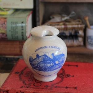 Może przedstawiać: Biały ceramiczny słoik z uchwytem, z niebieską ilustracją budynku i napisem "FORTNUM & MASON". Słoik stoi na czerwonej książce.