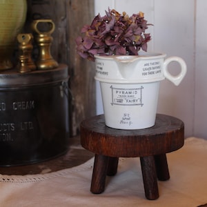 May include: A white ceramic pitcher with a handle and a spout, featuring black printed text that reads PYRAMID TRADE MARK FAIRY. The pitcher contains dried purple flowers and sits on a small, round, dark brown wooden stool with three legs.