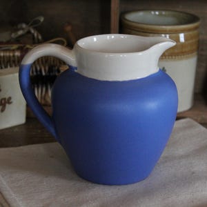 Może przedstawiać: Niebiesko-biały ceramiczny dzbanek z uchwytem. Dzbanek ma zaokrąglony korpus i dzióbek. Uchwyt jest biały z niebieskim akcentem. Inne ceramiczne pojemniki znajdują się w tle, wraz z pudełkiem fudge.