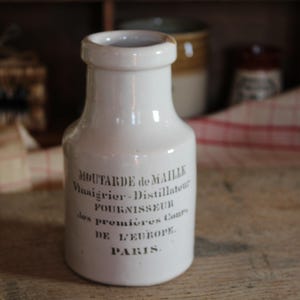 May include: A white ceramic bottle with a rounded body and a narrow neck. The bottle has black text that reads "Moutarde de Maille, Vinaigrier-Distillateur, Fournisseur des premières cours de l'Europe, PARIS."