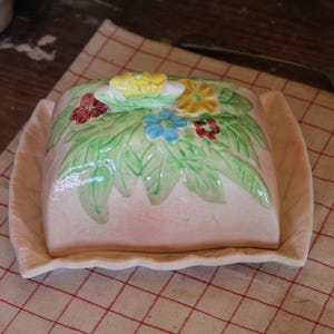 Könnte beinhalten: Eine Vintage-Butterdose aus rosa Keramik mit Deckel, verziert mit grünen Blättern und bunten Blumen. Die Schale steht auf einem passenden Tablett, perfekt zum Servieren von Butter. Die Butterdose ist eine charmante Ergänzung für jede Küche.
