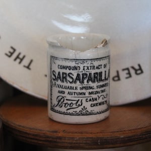 Könnte beinhalten: Ein kleines, antikes weißes Keramikgefäß mit der Aufschrift "Compound Extract of Sarsaparilla". Das Gefäß hat einen abgebrochenen Rand und steht auf einer Holzoberfläche. Das Etikett erwähnt auch "Boots Cash Chemists".