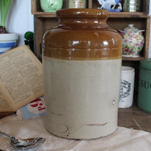 May include: A large, brown and white ceramic jar with a lid. The jar is sitting on a wooden table with a white cloth.