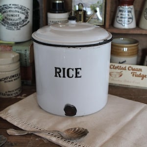 May include: A white enamel rice container with a lid and black trim. The word "RICE" is printed in black on the front. The container is part of a vintage kitchenware collection, with other jars and containers in the background.