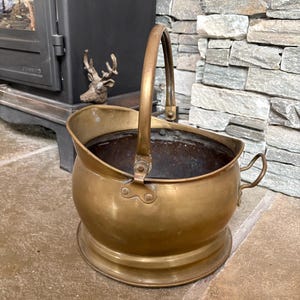May include: A round brass coal scuttle featuring a wide, open top, a tall arched handle attached with rivets, and a smaller side handle. The exterior has a polished metallic finish, while the interior is dark and deep with a circular base.