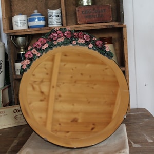 May include: A round wooden mirror with a decorative floral border of pink and green flowers. The mirror has a dark brown frame and is displayed on a wooden table. The background includes shelves with various vintage items.
