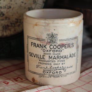 May include: A vintage, off-white ceramic jar of Frank Cooper's Seville Marmalade. The jar has a label with black text and a crest. The jar is on a red and white checkered cloth. The background includes a scale and a wooden object.