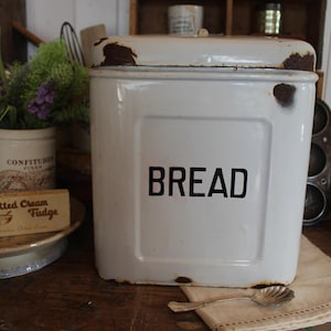 Può includere: Una scatola per il pane bianca con la scritta "BREAD" in nero. Il contenitore rettangolare ha un coperchio e angoli leggermente arrugginiti. Altri oggetti vintage da cucina sono sullo sfondo.