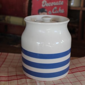 Peut inclure: Pot en céramique blanche avec un motif rayé bleu et blanc et un couvercle. Le pot est posé sur une nappe à carreaux rouge et blanc.
